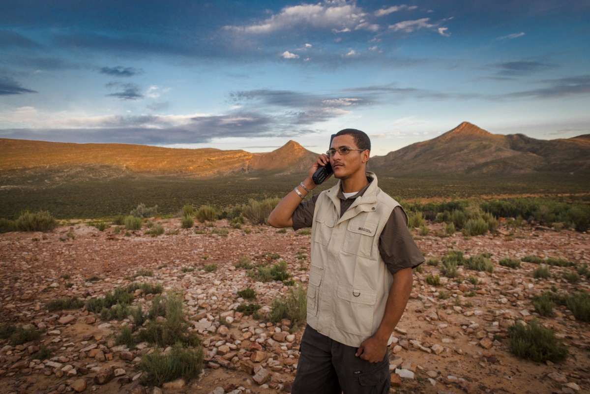 Man making a call on an Inmarsat IsatPhone 2 in dry scrubland below mountains